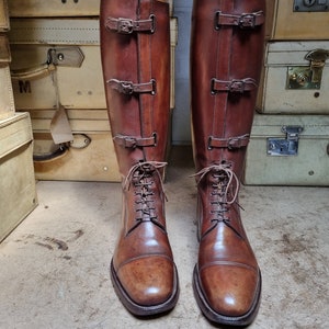 May include: A pair of brown leather riding boots with buckles and laces. The boots are on wooden boot trees and are displayed on a wooden surface.