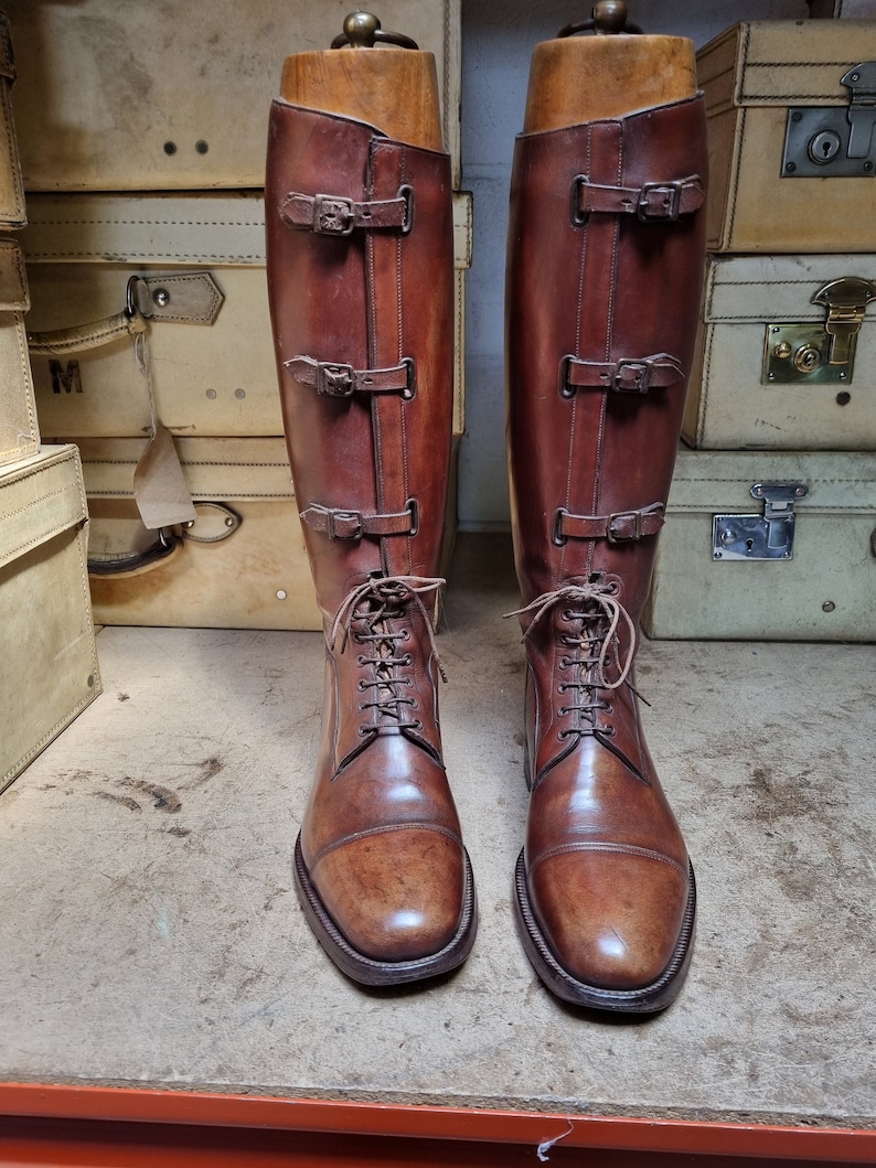 May include: A pair of brown leather riding boots with buckles and laces. The boots are on wooden boot trees and are displayed on a wooden surface.