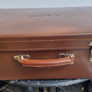 May include: A rectangular brown leather suitcase featuring a top handle, two silver-toned metal latches with keyholes, and visible stitching along the edges. The top surface of the suitcase is embossed with the text D. M. FORT in capital letters.