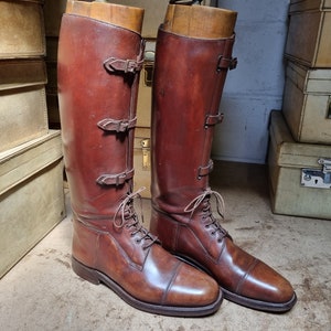 May include: A pair of brown leather riding boots with buckles and laces. The boots are on a wooden surface.