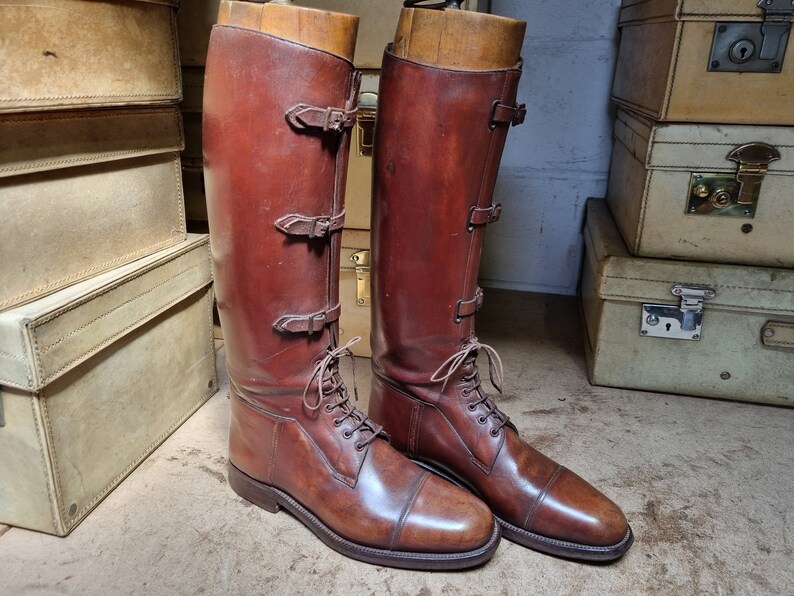 May include: A pair of brown leather riding boots with buckles and laces. The boots are on a wooden surface.