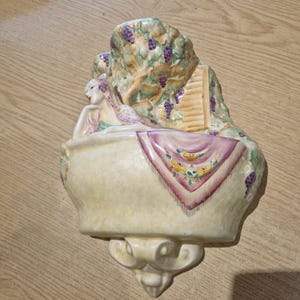May include: A ceramic wall pocket featuring a reclining figure resting on a pale yellow basin. The background depicts a tree with purple grapes and a small wooden structure. A pink and yellow patterned cloth drapes over the front of the basin.