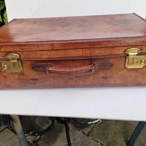 May include: A brown leather suitcase with brass fittings. The suitcase features a handle and two brass clasps. The leather has a worn look, indicating age and use. The rectangular suitcase appears to be in good condition, with visible stitching.