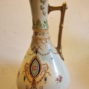 May include: A cream-colored ceramic pitcher with a gold-toned handle, featuring a crackled glaze finish. The body is decorated with intricate floral motifs, blue and gold filigree patterns, and green leaf accents near the flared spout.