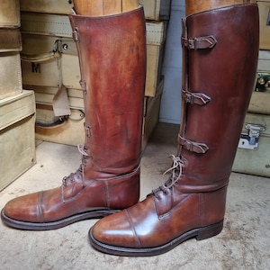 May include: A pair of brown leather riding boots with buckles and laces. The boots are on wooden forms and are displayed against a background of vintage suitcases.