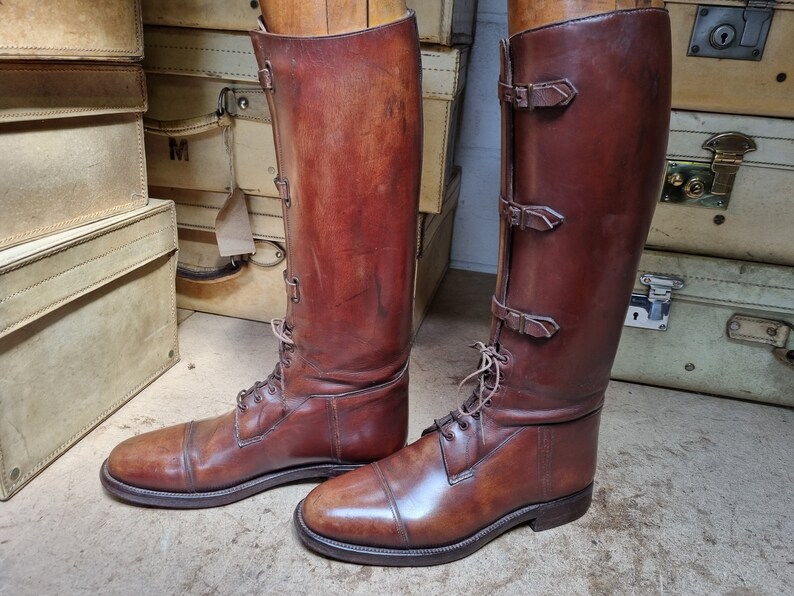 May include: A pair of brown leather riding boots with buckles and laces. The boots are on wooden forms and are displayed against a background of vintage suitcases.