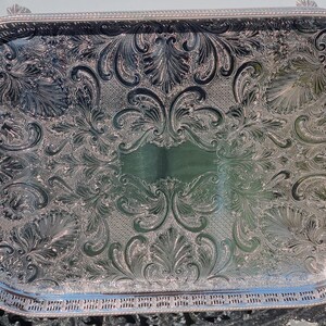 May include: A rectangular silver-toned serving tray featuring an intricate embossed floral and scrollwork pattern across its surface. The tray has a raised, pierced gallery border and two curved handles on the sides, with a polished reflective finish.