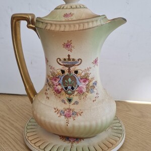 May include: A ceramic coffee pot with a lid and matching base, featuring a cream-colored body with green-tinted edges, a gold-painted handle, and a central decorative motif of pink roses, blue accents, and a small urn illustration.