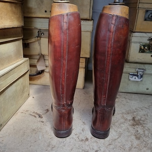 May include: A pair of brown leather riding boots with a tall shaft and a pointed toe. The boots are standing on a light brown surface.