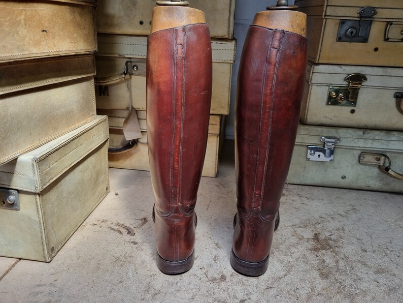 May include: A pair of brown leather riding boots with a tall shaft and a pointed toe. The boots are standing on a light brown surface.