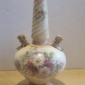 May include: A cream-colored ceramic vase with a bulbous body and a tall, spiraled neck. The vase features a floral pattern in shades of pink, white, and green, with two ornate, gold-toned handles on the sides and a decorative base.