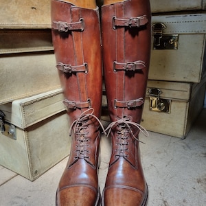 May include: A pair of brown leather riding boots with laces and buckles. The boots are on wooden shoe trees and are sitting on top of a stack of suitcases.