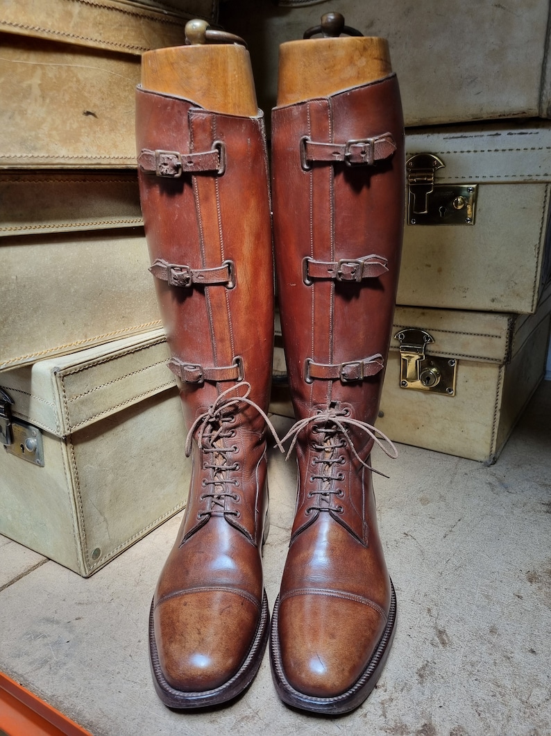May include: A pair of brown leather riding boots with laces and buckles. The boots are on wooden shoe trees and are sitting on top of a stack of suitcases.