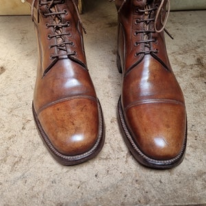 May include: A pair of brown leather lace-up boots with a cap toe design. The boots have a polished finish and are made of high-quality leather.