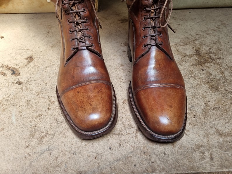 May include: A pair of brown leather lace-up boots with a cap toe design. The boots have a polished finish and are made of high-quality leather.