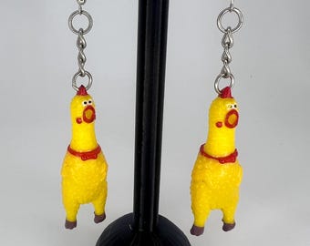 May include: A pair of novelty earrings featuring yellow rubber chicken charms. Each chicken has a red comb, bow tie, and open beak. The earrings hang from silver-colored fish hook ear wires and chain links.