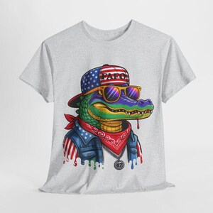 May include: A light gray t-shirt featuring a cartoon alligator wearing sunglasses, a bandana, and a denim jacket with an American flag design. The alligator is wearing a hat with the text "PAF0".