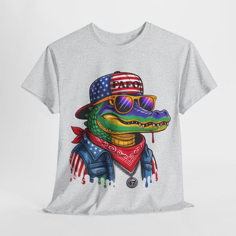 May include: A light gray t-shirt featuring a cartoon alligator wearing sunglasses, a bandana, and a denim jacket with an American flag design. The alligator is wearing a hat with the text "PAF0".