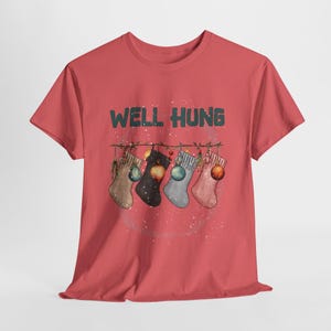 May include: Red t-shirt with the text "WELL HUNG" in green letters. Below the text are five Christmas stockings hanging on a string, each with a different color and design, and decorated with ornaments.