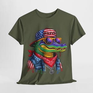 May include: Olive green t-shirt featuring a cartoon alligator wearing a patriotic hat, sunglasses, and a bandana. The alligator has a denim jacket with American flag accents and a silver pendant. The hat says "PAF0".