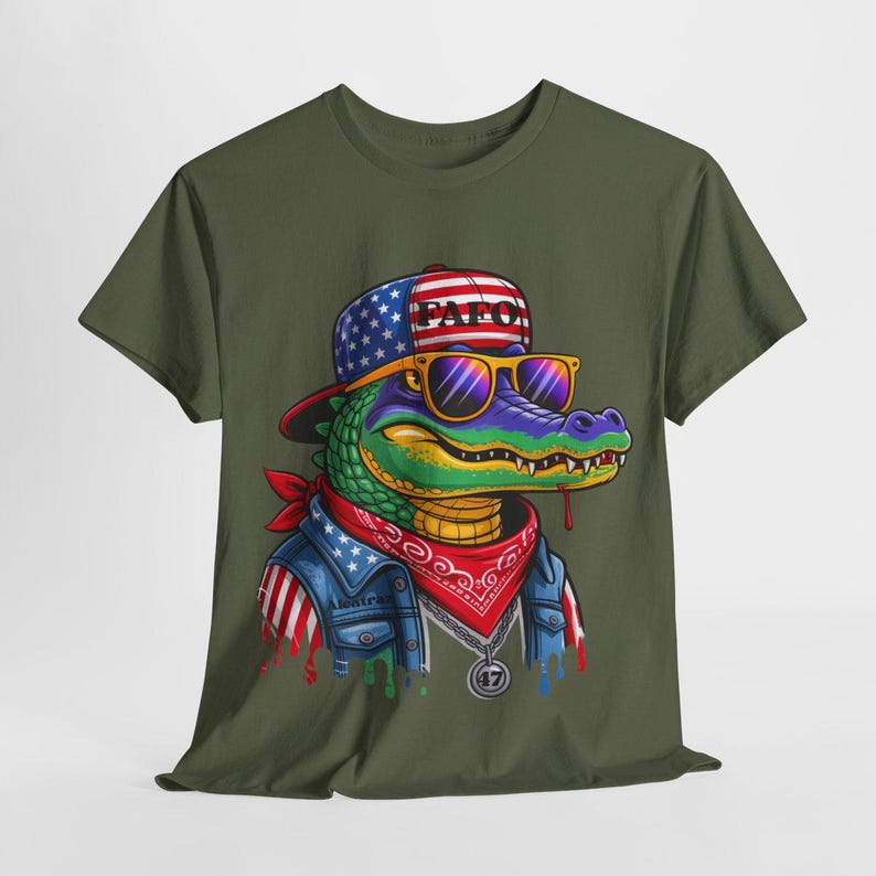 May include: Olive green t-shirt featuring a cartoon alligator wearing a patriotic hat, sunglasses, and a bandana. The alligator has a denim jacket with American flag accents and a silver pendant. The hat says "PAF0".