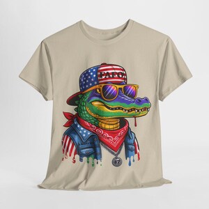 May include: Tan t-shirt featuring a cartoon alligator wearing a patriotic hat, sunglasses, and a bandana. The alligator has a denim jacket with American flag accents. The hat says "PAF0". The alligator has a pendant that says "47".