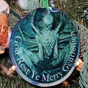 Op de afbeelding: Een ronde, blauwe en groene kerstbal met een glinsterende afwerking. De bal heeft een zwart-wit afbeelding van een wezen met de tekst "God Rest Ye Merry Gentlemen".