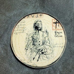 May include: A round, cream-coloured coaster with a black and white illustration of a skeletal figure holding a paintbrush and palette. The coaster has a gold glitter finish and the text "Games & Gissars" in black lettering.