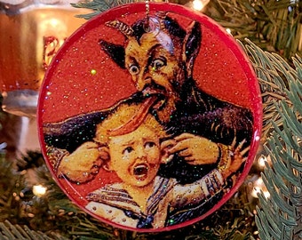 Puede incluir: Un adorno navideño rojo con purpurina que representa a un diablo de dibujos animados con cuernos y la lengua fuera, sujetando la oreja de un niño. El niño lleva una camisa de marinero y tiene una expresión de miedo.