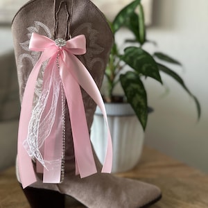 Op de afbeelding: Een lichtbruine su&egrave;de cowboylaars versierd met een decoratief accessoire bestaande uit een roze satijnen strik, een centrale strass-versiering, hangende roze satijnen linten, een strook witte kant en een verticale streng met strass-steentjes.
