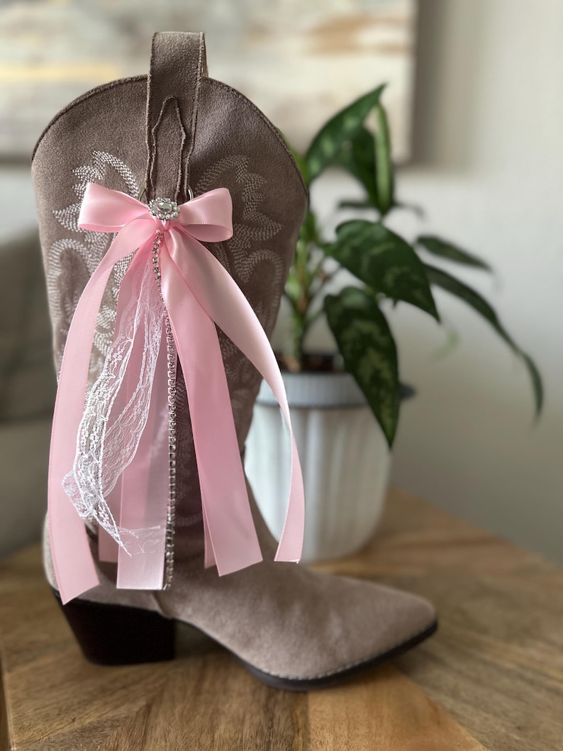 Op de afbeelding: Een lichtbruine su&egrave;de cowboylaars versierd met een decoratief accessoire bestaande uit een roze satijnen strik, een centrale strass-versiering, hangende roze satijnen linten, een strook witte kant en een verticale streng met strass-steentjes.