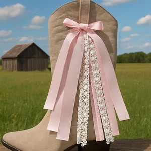 May include: A beige suede cowboy boot decorated with a pink satin bow and ribbons. White lace trim adds detail. The boot is set against a backdrop of a rural landscape with a wooden barn.
