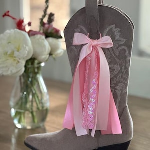 Op de afbeelding: Een taupekleurige suède cowboylaars met ingewikkelde ton-sur-ton stiksels op de schacht. Aan de zijkant is een decoratief accessoire bevestigd, bestaande uit een lichtroze satijnen strik met neerhangende linten en een streng glinsterende roze lovertjes.