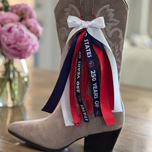 May include: A tan suede cowboy boot featuring white decorative stitching and a cluster of ribbons attached to the side. The ribbons are white, red, and navy blue, with the navy blue ribbon printed with the text 250 YEARS OF STATES.