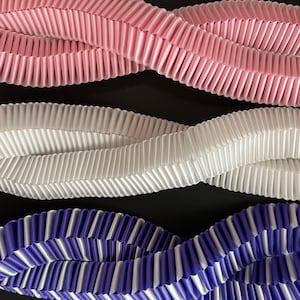 May include: Close-up of three wavy, pleated ribbons in pink, white, and blue with white stripes. The ribbons are arranged against a black background, creating a visually appealing contrast. The pleated texture adds depth.
