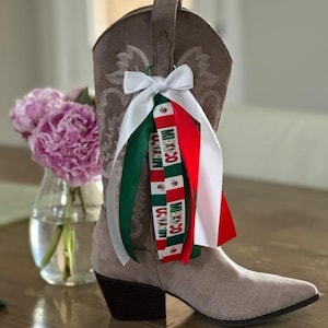 May include: A taupe suede cowboy boot featuring decorative stitching and a cluster of ribbons tied to the side. The ribbons include solid white, green, and red satin strips alongside a patterned ribbon with the text MEXICO and small emblems.