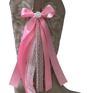 Op de afbeelding: Een beige su&egrave;de cowboylaars met een decoratief accessoire aan de zijkant. Het accessoire bestaat uit een roze satijnen strik, een centraal rond strass-ornament en verschillende hangende linten van roze satijn en witte kanten stof.