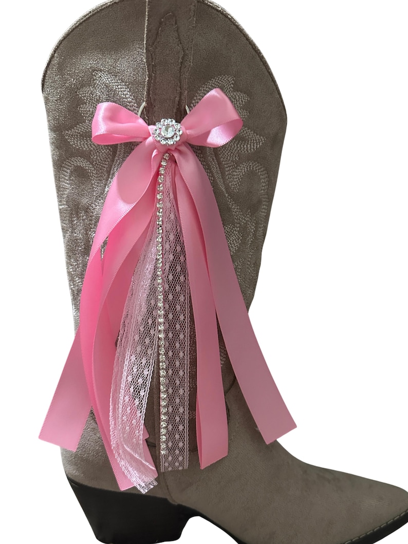Op de afbeelding: Een beige su&egrave;de cowboylaars met een decoratief accessoire aan de zijkant. Het accessoire bestaat uit een roze satijnen strik, een centraal rond strass-ornament en verschillende hangende linten van roze satijn en witte kanten stof.