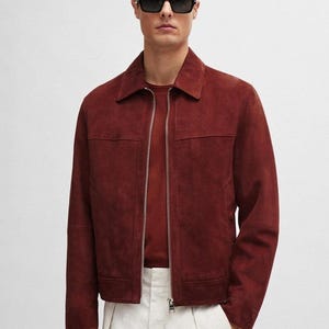 Puede incluir: Chaqueta de ante burdeos con cierre de cremallera, usada sobre un suéter burdeos a juego. La chaqueta tiene un cuello clásico y se combina con pantalones blancos. La persona lleva gafas de sol negras.