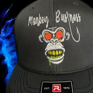 May include: Black and white embroidered trucker hat with a monkey face with red and orange eyes and a green drool. The text "Monkey Business" is embroidered on the front of the hat. The hat has a black mesh back and a black bill with a red "R" logo and the text "EST. 1970".