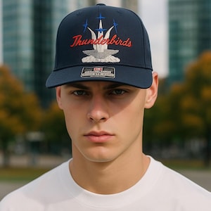 May include: Navy blue baseball cap with the word "Thunderbirds" embroidered in red script. The cap features a stylized emblem of a bird and three blue lines. The cap has a flat brim and a structured fit.