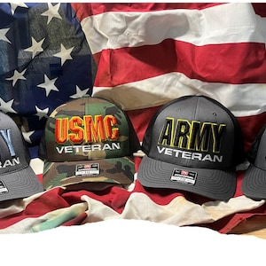 May include: Four black mesh trucker hats with different branch of service logos and the word "Veteran" embroidered on each. The logos are Navy, USMC, Army, and USAF. The hats are arranged in a row on a red, white, and blue American flag.