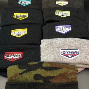 May include: A stack of ten black knit beanies with different colored embroidered patches that say "B-52 Bomber Pride". The patches have different colored stars and outlines.