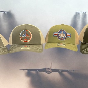 May include: Four green and beige trucker hats with different embroidered designs featuring American flags and airplanes. The designs include a peace sign, a star with wings, and a plane with the words "Peace Through Strength".