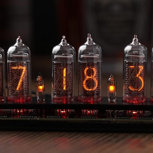 Puede incluir: Un reloj digital con seis tubos de vidrio que muestran la hora 17:34. Los números brillan en naranja sobre un fondo oscuro. El reloj está sobre una base negra con componentes electrónicos visibles.