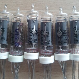 Może przedstawiać: Sześć przezroczystych szklanych lamp pr&oacute;żniowych z srebrnymi drutami i białymi czapkami. Każda lampa ma czarną etykietę z tekstem "ИН-16" i numerem, takim jak "9102" lub "9105".