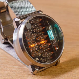 K&ouml;nnte beinhalten: Eine runde Armbanduhr mit silberfarbenem Metall-Mesh-Armband und rundem Geh&auml;use. Das Zifferblatt hat einen schwarzen Hintergrund mit mehreren kleinen Glas-Vakuumr&ouml;hren, von denen eine hellorange und eine t&uuml;rkis leuchtet, sowie r&ouml;mischen Ziffern.