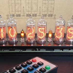 Op de afbeelding: Een digitale klok met zes glazen buizen die de tijd 10:25:35 weergeven. De buizen gloeien met een oranje licht, en de klok staat op een zwarte basis. Een afstandsbediening staat op de voorgrond.