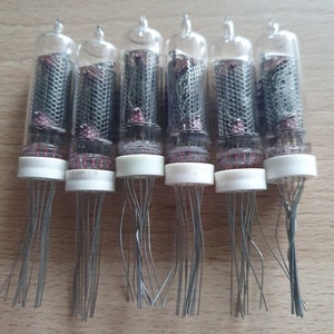 Może przedstawiać: Sześć zabytkowych szklanych lamp pr&oacute;żniowych z metalowymi siatkami i białymi ceramicznymi podstawami. Każda lampa ma wiele cienkich metalowych przewod&oacute;w wychodzących z podstawy.