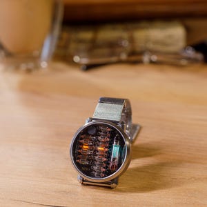 K&ouml;nnte beinhalten: Eine runde Armbanduhr mit einem silberfarbenen Metall-Mesh-Armband und einem dunklen, kreisf&ouml;rmigen Zifferblatt. Das Zifferblatt zeigt freiliegende elektronische Komponenten, darunter mehrere kleine Glasr&ouml;hren mit leuchtend orangefarbenen und gr&uuml;nen Elementen. Das Wort NIWA ist in Wei&szlig; auf der unteren linken Seite des Zifferblatts aufgedruckt.